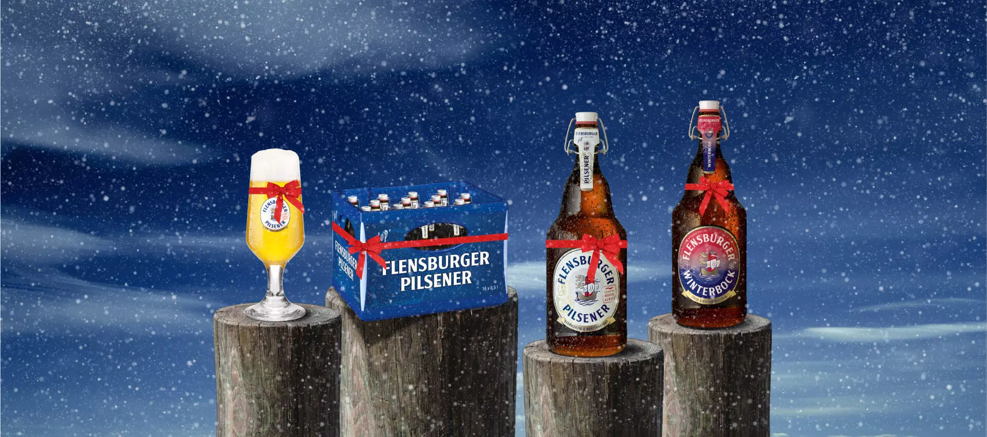 Bierglas, Mega-Plops von FLENS und eine Kiste Bier auf Holzpollern im Schnee und mit Geschenkeband.