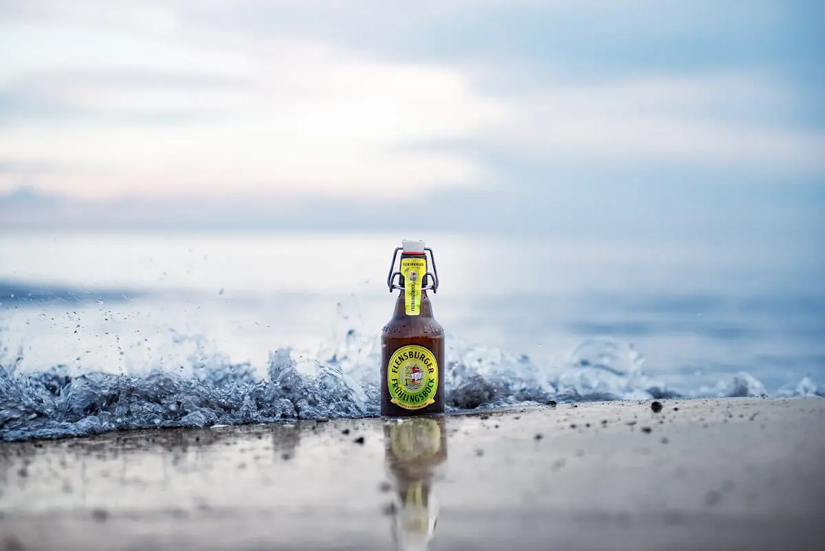 Flensburger-Frühlingsbock-Bierflasche am Strand, Wellenbrandung und Spiegelung im nassen Sand.