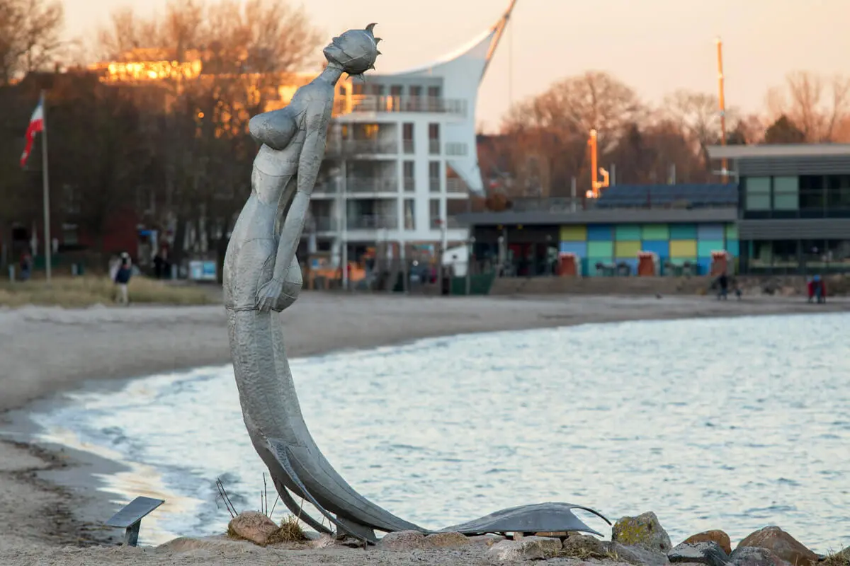 Metallene Meerjungfrau-Skulptur am Strand vor Promenade von Eckernförde und ruhiger Ostsee im Abendlicht.