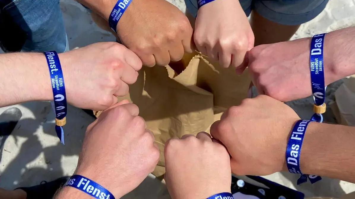 Gruppe von Personen bildet einen Kreis und zeigt jeweils ihr blaues FLENS StrandGut Armband einem Sandstrand.