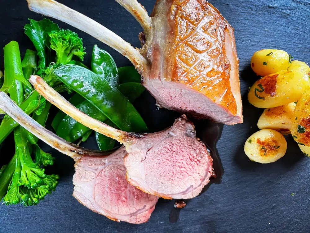 Medium-rare Lammkoteletts mit Brokkolini, Zuckerschoten und gebratenen Mini-Kartoffeln auf Schieferplatte.