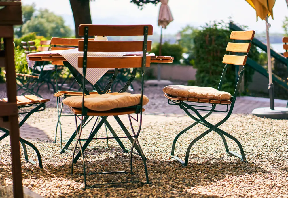 Sonniger Biergarten mit hölzernen Bistrostühlen und Sitzkissen auf Kies.