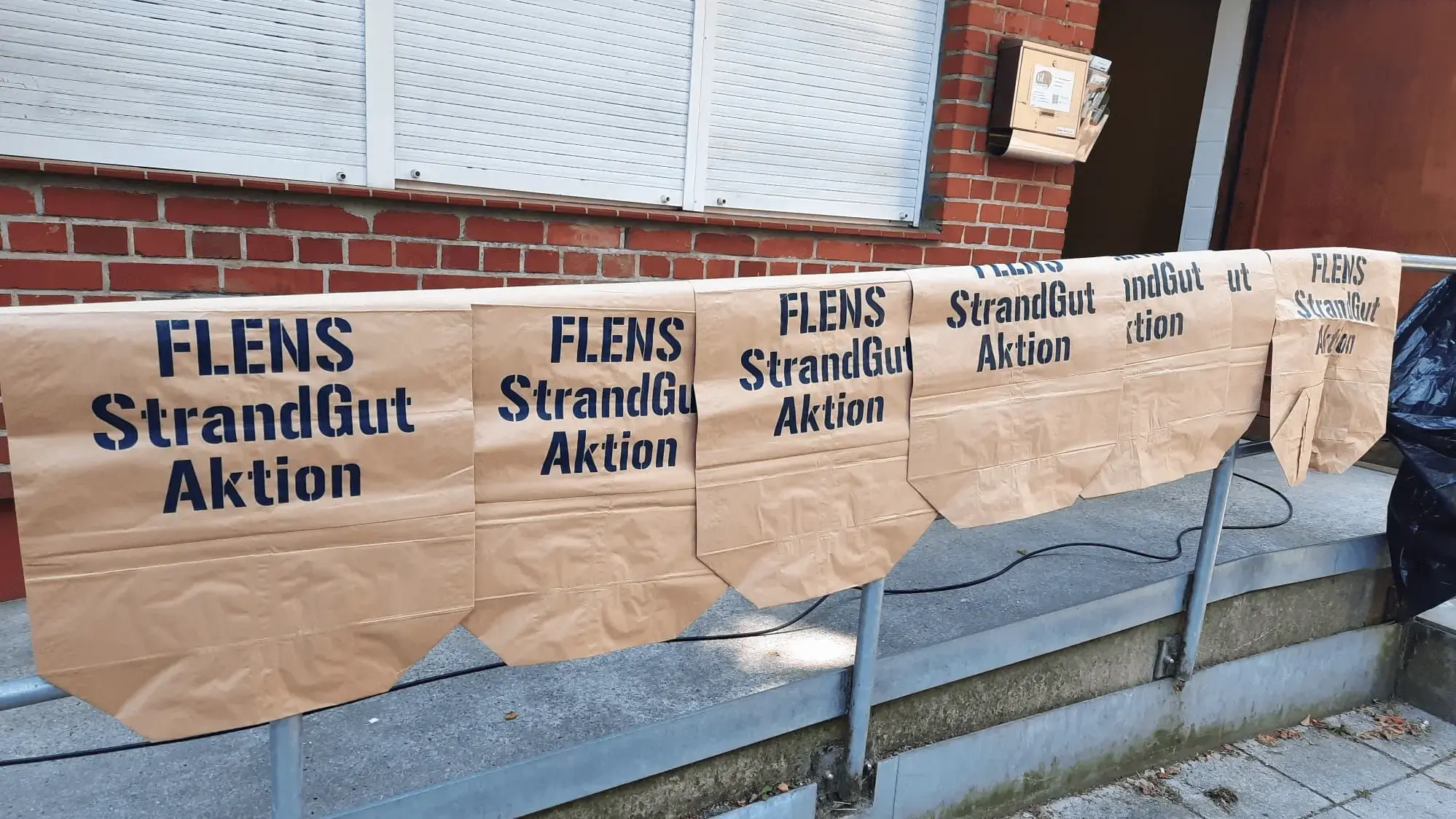 Papiermülltüten mit Aufschrift "FLENS StrandGut Aktion" vor einem Ziegelgebäude aufgehängt.