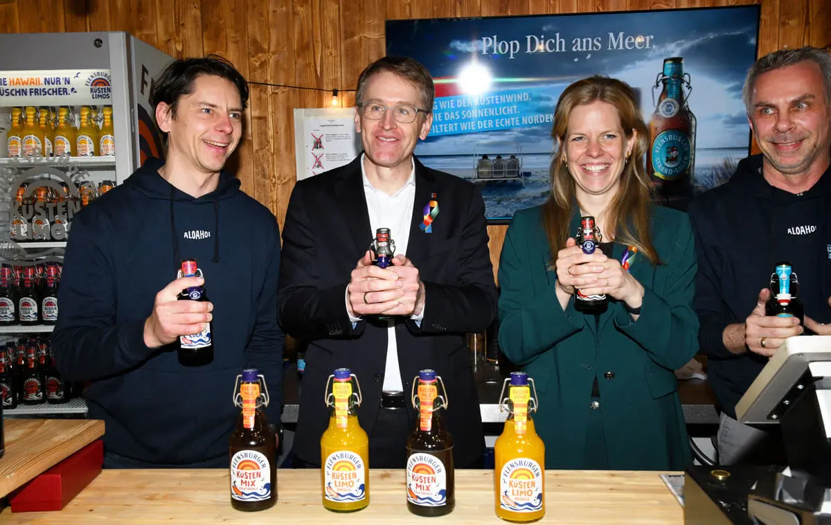 Vier Personen, darunter Ministerpräsident Daniel Günther, hinterm Tresen am Messestand und halten Flensburger Küstenlimo-Flaschen; Hintergrundbanner mit Plop Dich ans Meer.