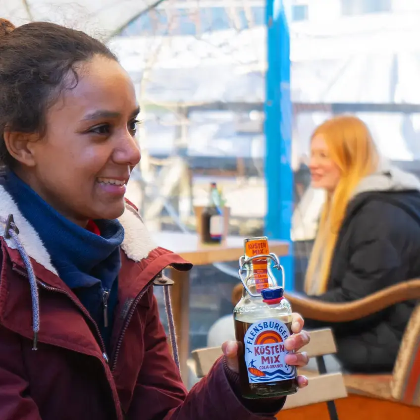 Zwei Personen stoßen mit Flensburger Küstenmix-Flaschen an im winterlichen Außencafé.