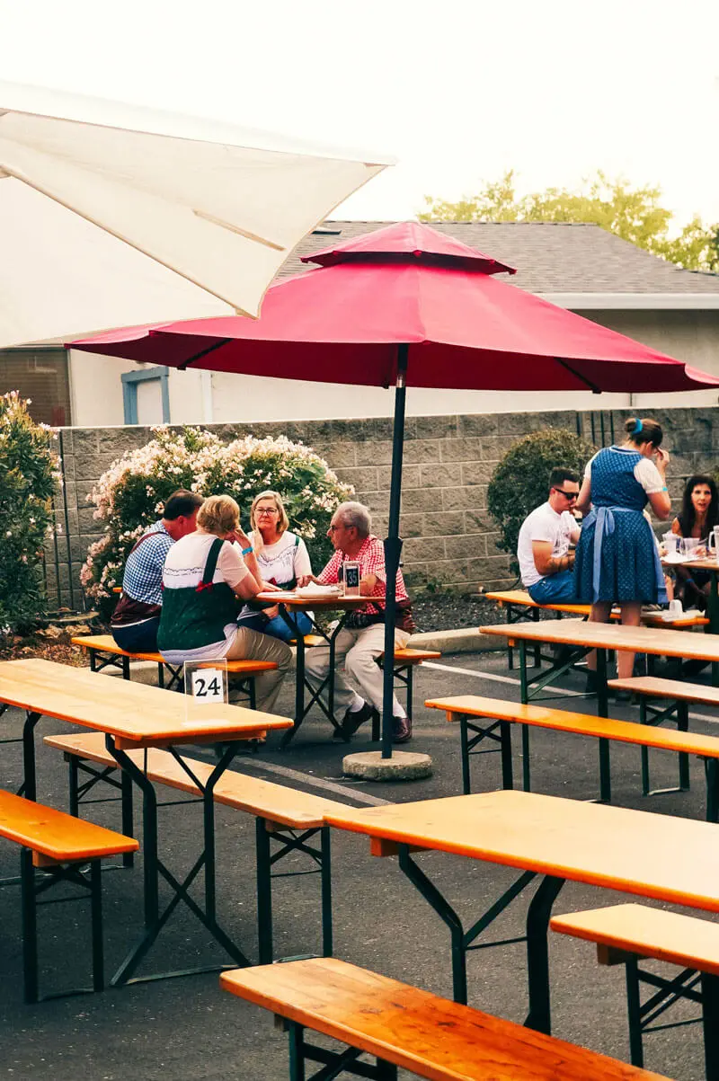 Biergarten im Freien mit Holzbänken, roten Sonnenschirmen und Gästen beim Essen und Trinken.