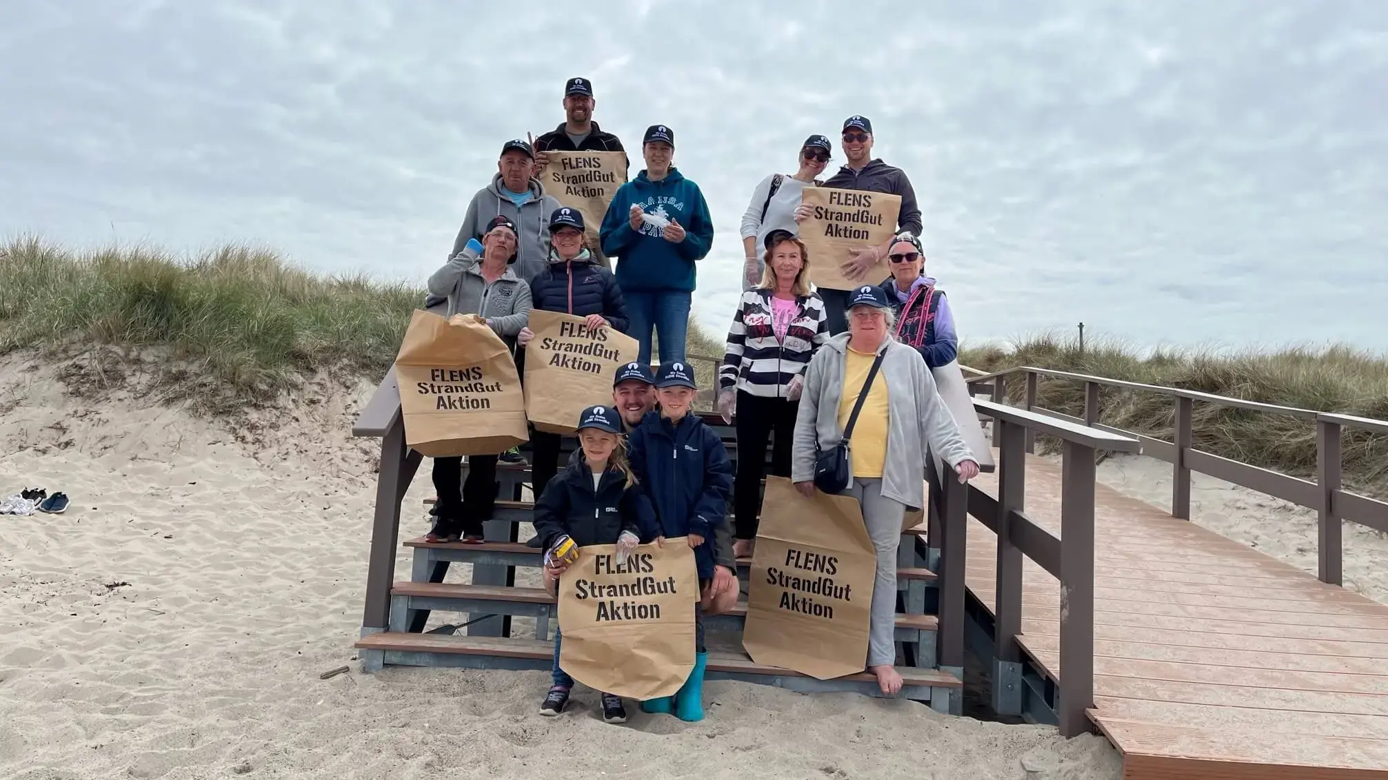 Gruppe von Freiwilligen mit Säcken bei FLENS Strandreinigungsaktion auf einer Holztreppe.