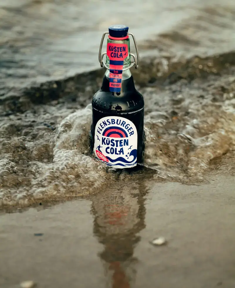 Flensburger-Küstencola-Bügelverschlussflasche in der Brandung am Sandstrand.