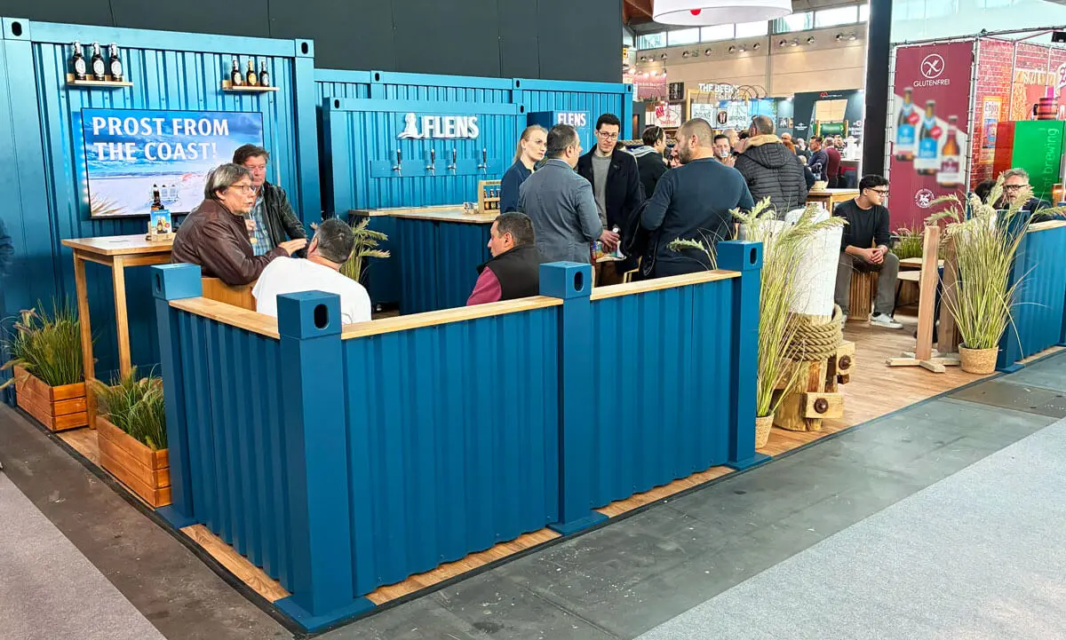 Blauer Messestand aus Metallpaneelen mit Barbereich, Besuchergruppen, Gräserpflanzen und Bierverkostung.