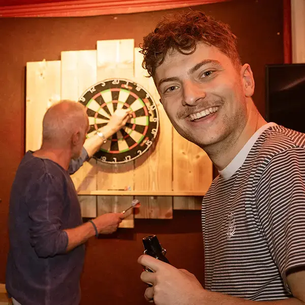 Freunde spielen Darts an einer Dartscheibe in einer gemütlichen Bar und trinken FLENS, Frau lacht in die Kamera.