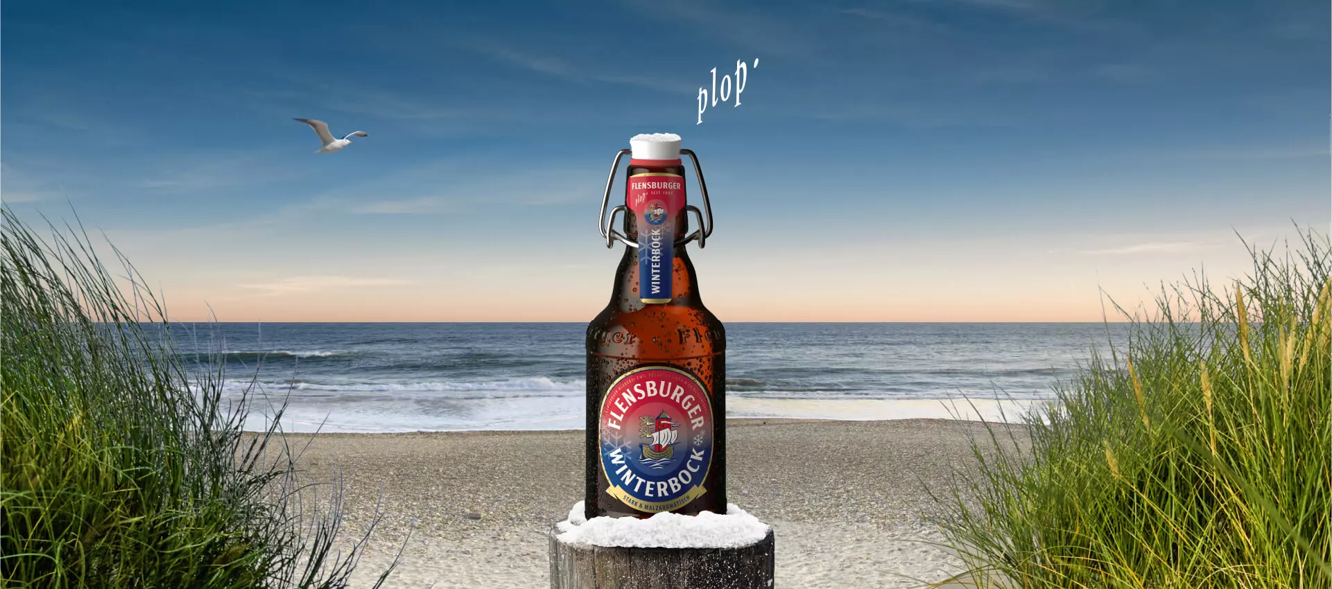 Eine Flasche Flensburger Winterbock auf einem Poller mit Schnee vor einer Strandlandschaft.