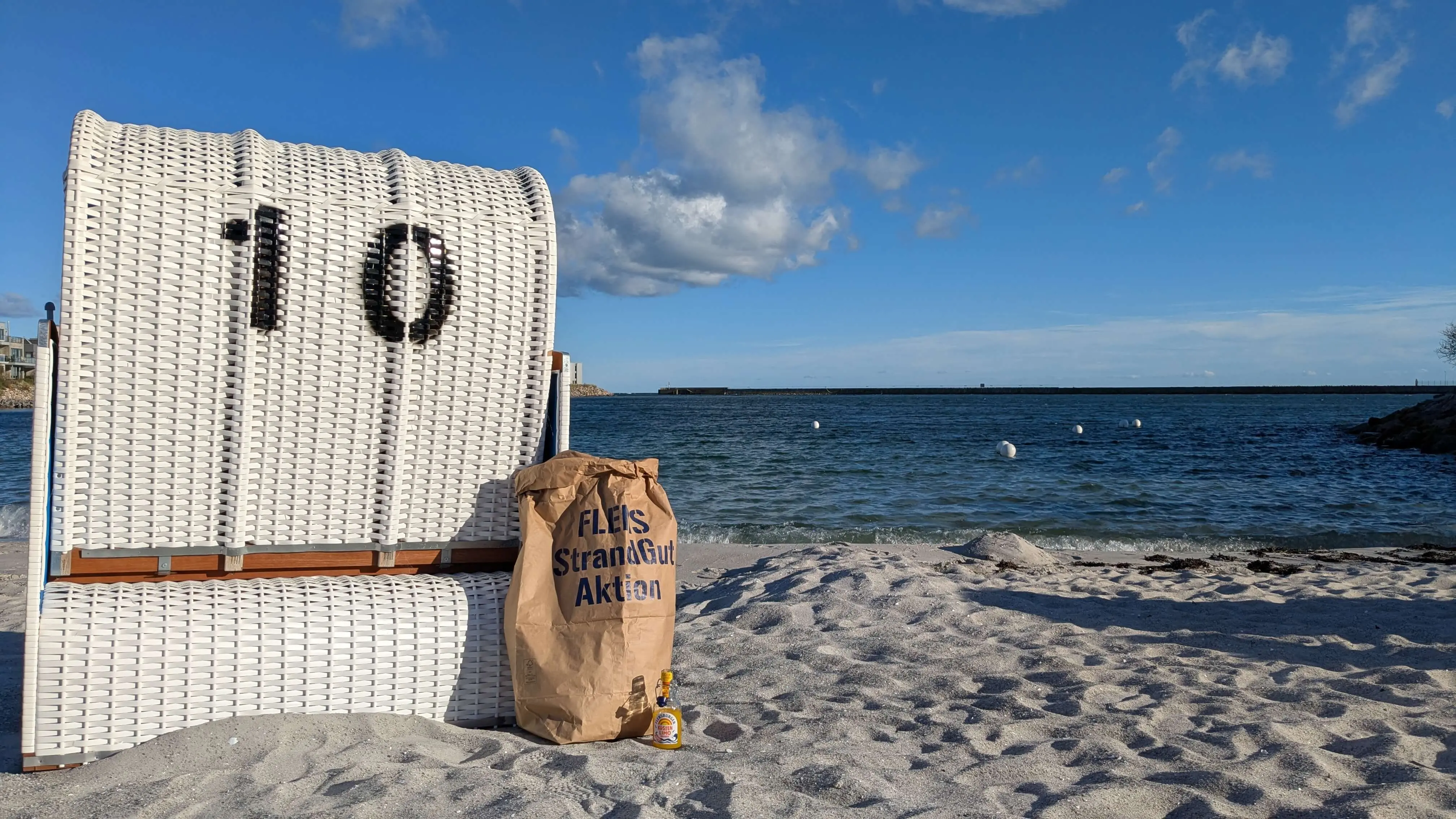 Strandkorb Nummer 10 am Sandstrand mit Müllsack der FLENS StrandGut Aktion und FLENS Flasche, Meerblick und Wolkenhimmel.