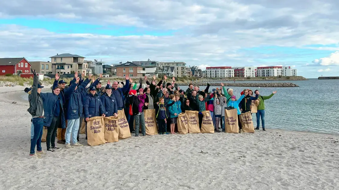 Menschengruppe mit FLENS StrandGut-Müllsammeltüten an der Strand-Promenade von Olpenitz, alle jubeln und heben die Arme.