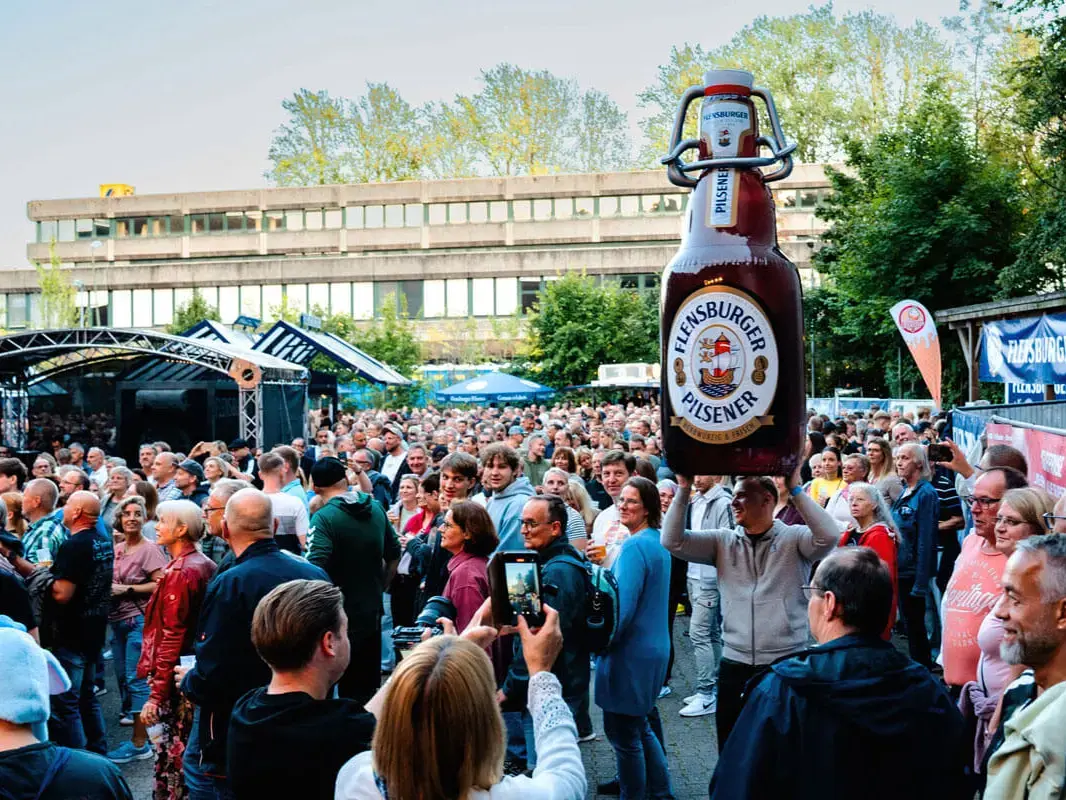 FLENS Festival mit großer Menschenmenge, Bühne und übergroßer, aufblasbarer Flensburger-Pilsener-Flasche.