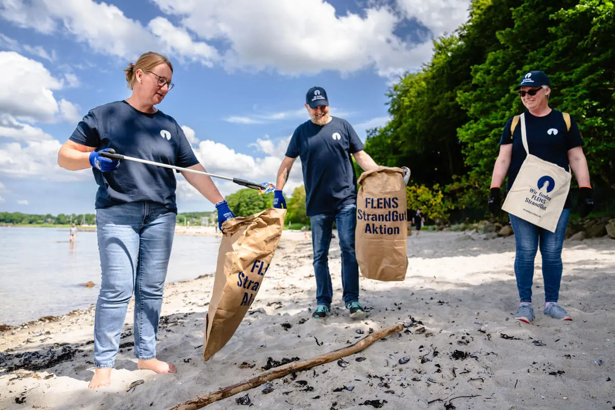 Freiwillige bei einer FLENS Strandreinigung mit Müllsäcken und Greifzangen an einem sonnigen Tag.