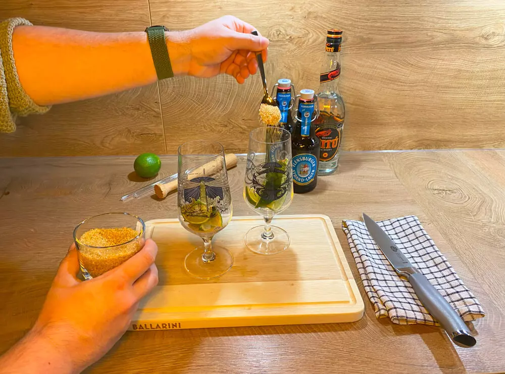 Ein Teelöffel brauner Rohrzucker wird in ein Bierglas eingefüllt, dahinter Cocktailutensilien.