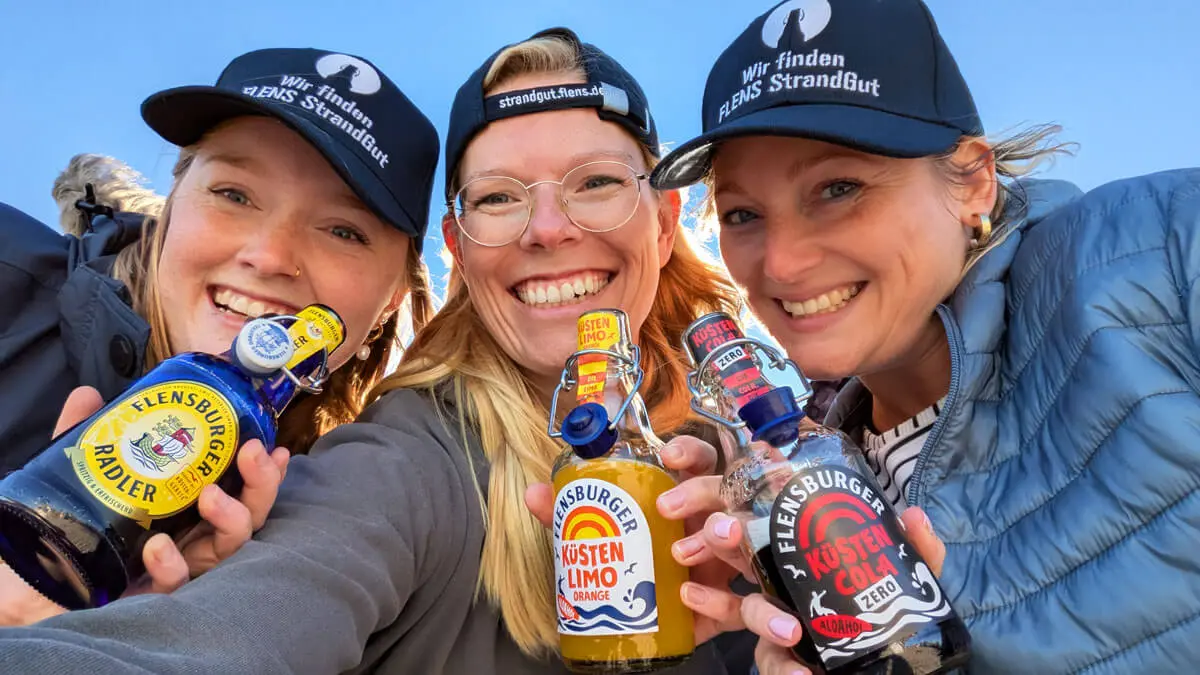 Drei lächelnde Frauen mit Flensburger-Flaschen (Radler, Küstenlimo Orange, Küstencola Zero), Strandgut-Kappen im Freien.