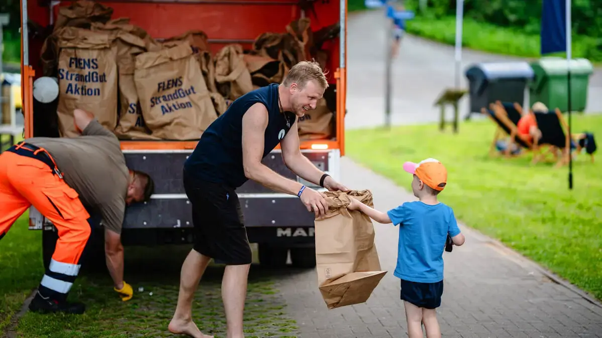 Freiwilliger nimmt Müllsack von einem Kind bei einer FLENS StrandGut Aktion entgegen.