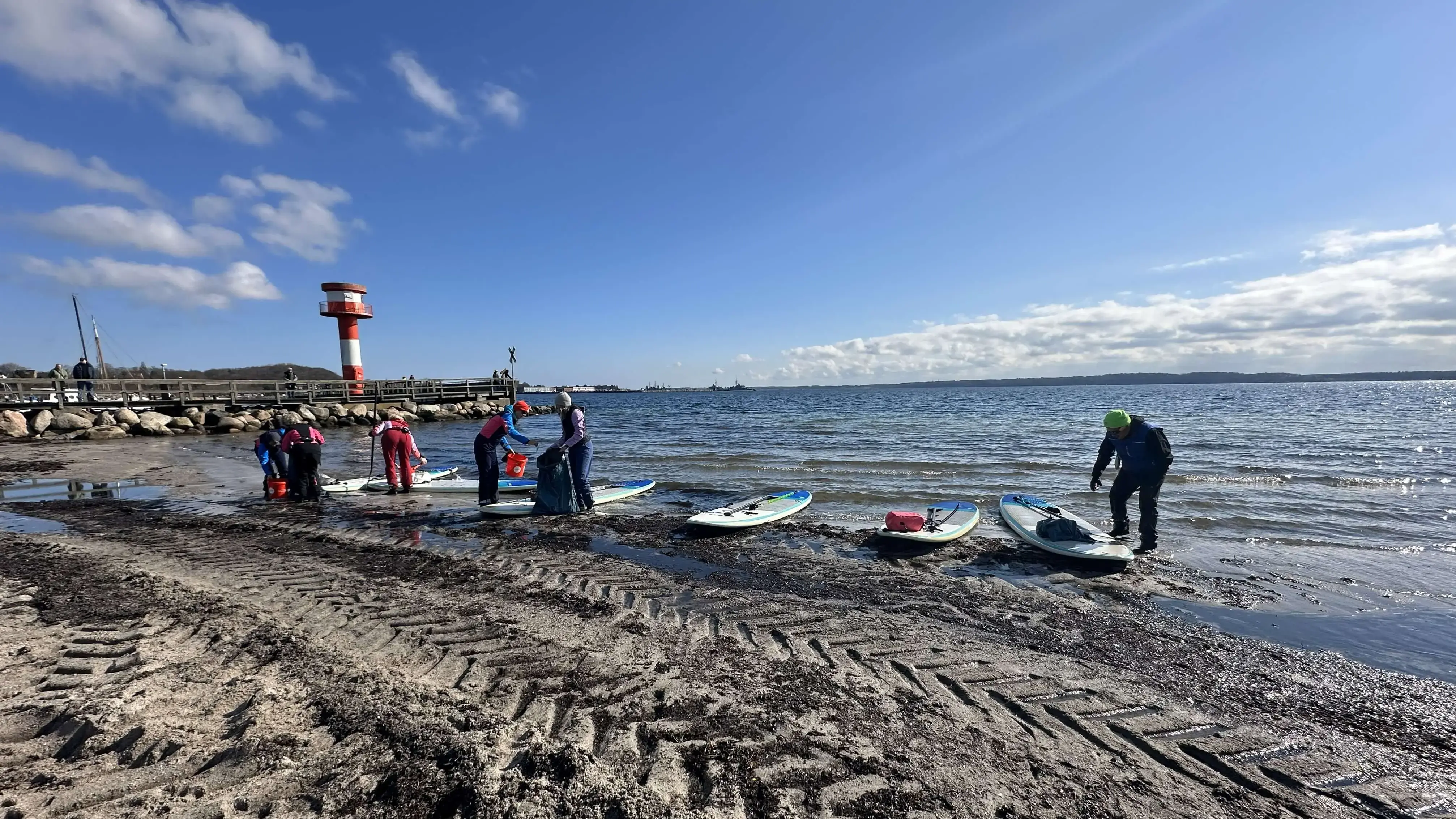 Menschen bereiten Paddleboards am sandigen Ufer mit Leuchtturm im Hintergrund vor, um damit auf dem Wasser Müll für die FLENS StrandGut Aktion zu sammeln.