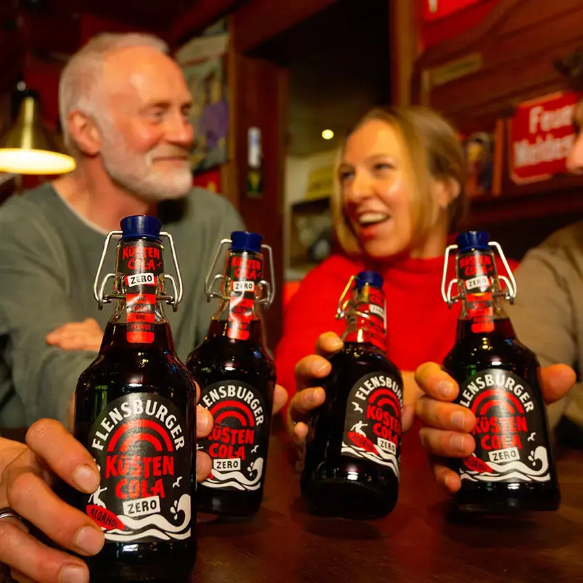 Vier Freunde in Kneipe stoßen mit Flensburger Küstencola-Zero-Flaschen an, gemütliche Bar-Atmosphäre.