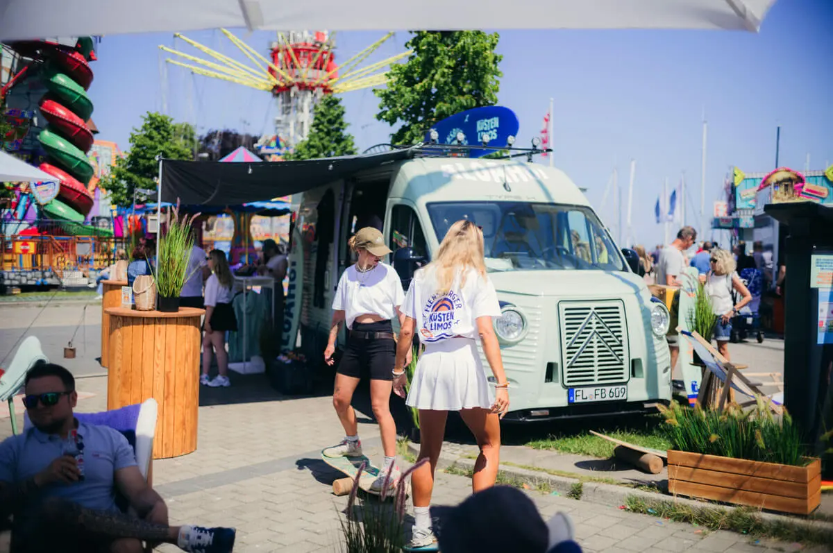 Sommerfestival am Hafen mit Vintage-Van, Promostand, Besucher und buntem Jahrmarkt im Hintergrund.