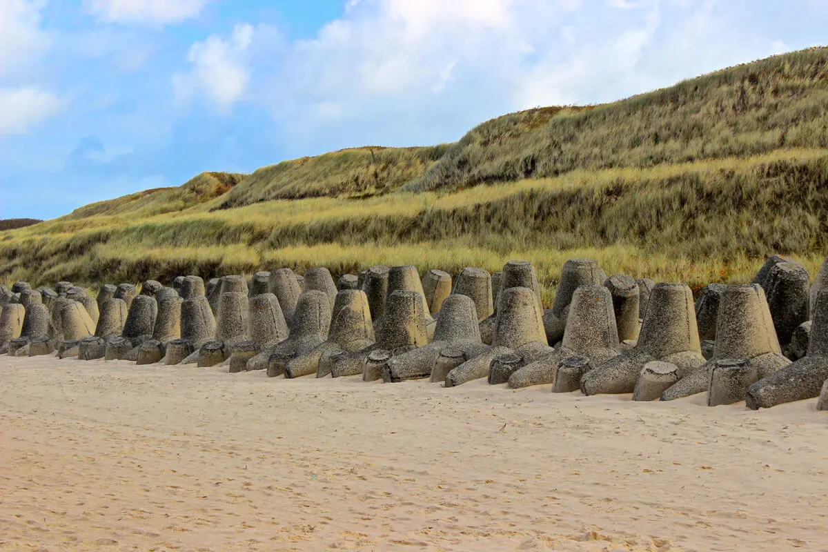 Sandstrand auf Sylt mit Beton-Tetrapoden als Wellenbrecher vor grasbewachsenen Dünen und blauem Himmel.