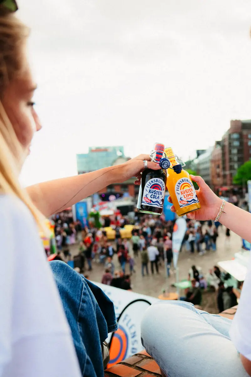 Zwei Personen stoßen mit Flensburger Küstencola und Küstenlimo an am Hamburger Fischmarkt mit Menschenmenge und Elbphilharmonie im Hintergrund.