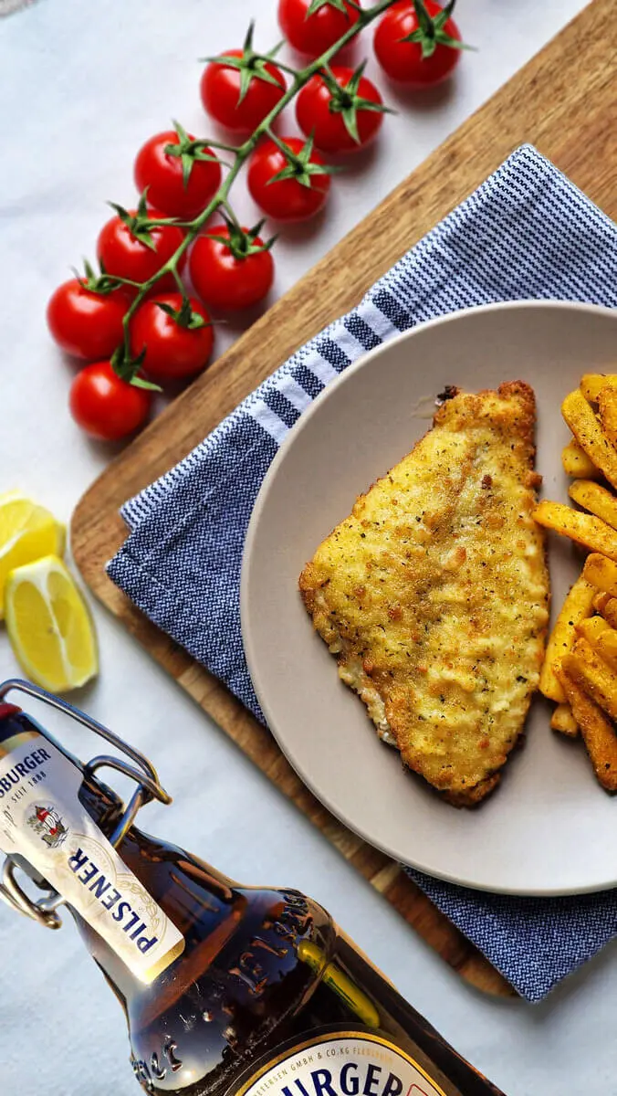 Knuspriger Backfisch im Bierteig mit Pommes, Kirschtomaten, Zitronenscheiben und Bierflasche auf Holzbrett.