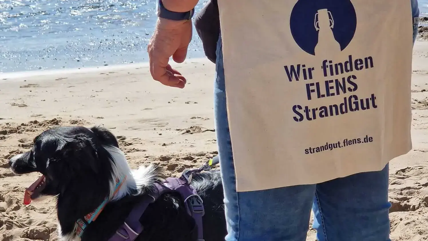 Person mit "FLENS Strandgut"-Stofftasche und Hund am Strand.