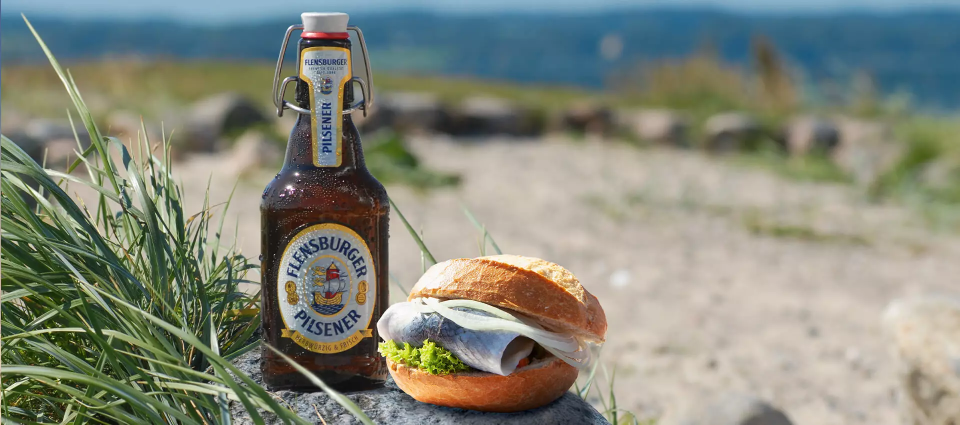 Flensburger Pilsener mit einem Fischbrötchen am Strand.