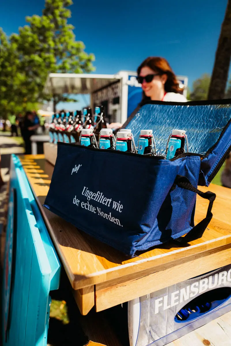 Flensburger Bierflaschen in blauer Kühltasche auf Holztresen bei sonnigem Outdoor-Event.