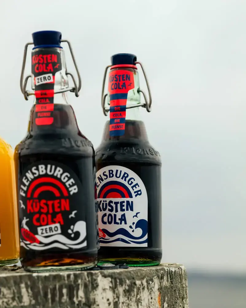Zwei Flaschen Küstencola von Flensburger mit Bügelverschluss auf verwittertem Pfahl vor Meereshorizont.