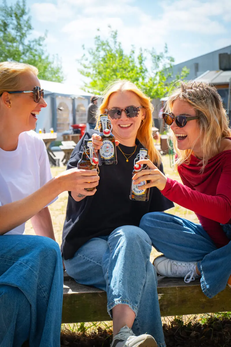 Drei Frauen stoßen mit Bierflaschen an im sonnigen Biergarten, entspannt in Jeans.