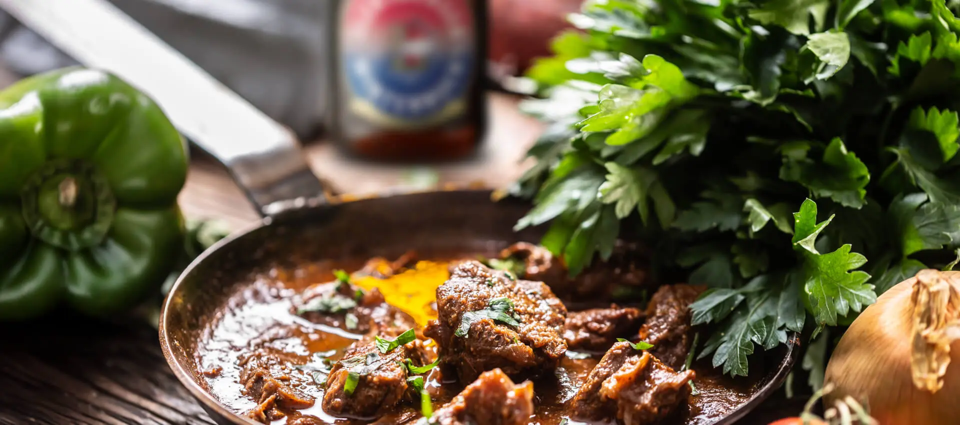 Appetitliches Rinderbiergulasch in einer Pfanne mit frischer Petersilie und Gemüse, im Hintergrund eine Flasche Flensburger Winterbock.