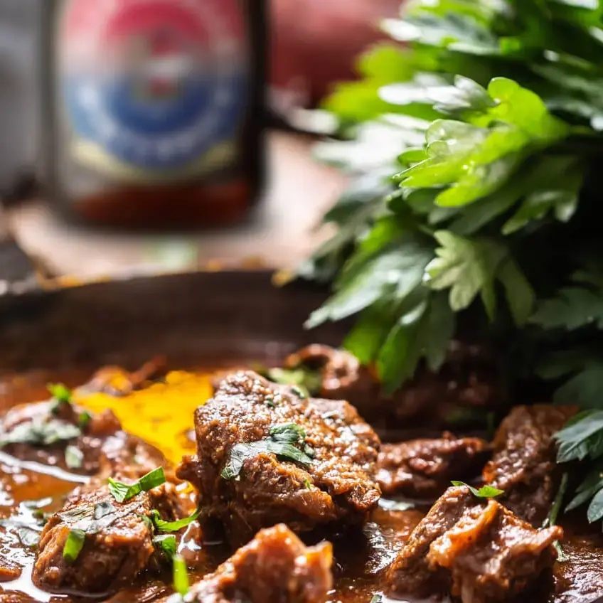 Appetitliches Rinderbiergulasch in einer Pfanne mit frischer Petersilie und Gemüse, im Hintergrund eine Flasche Flensburger Winterbock.