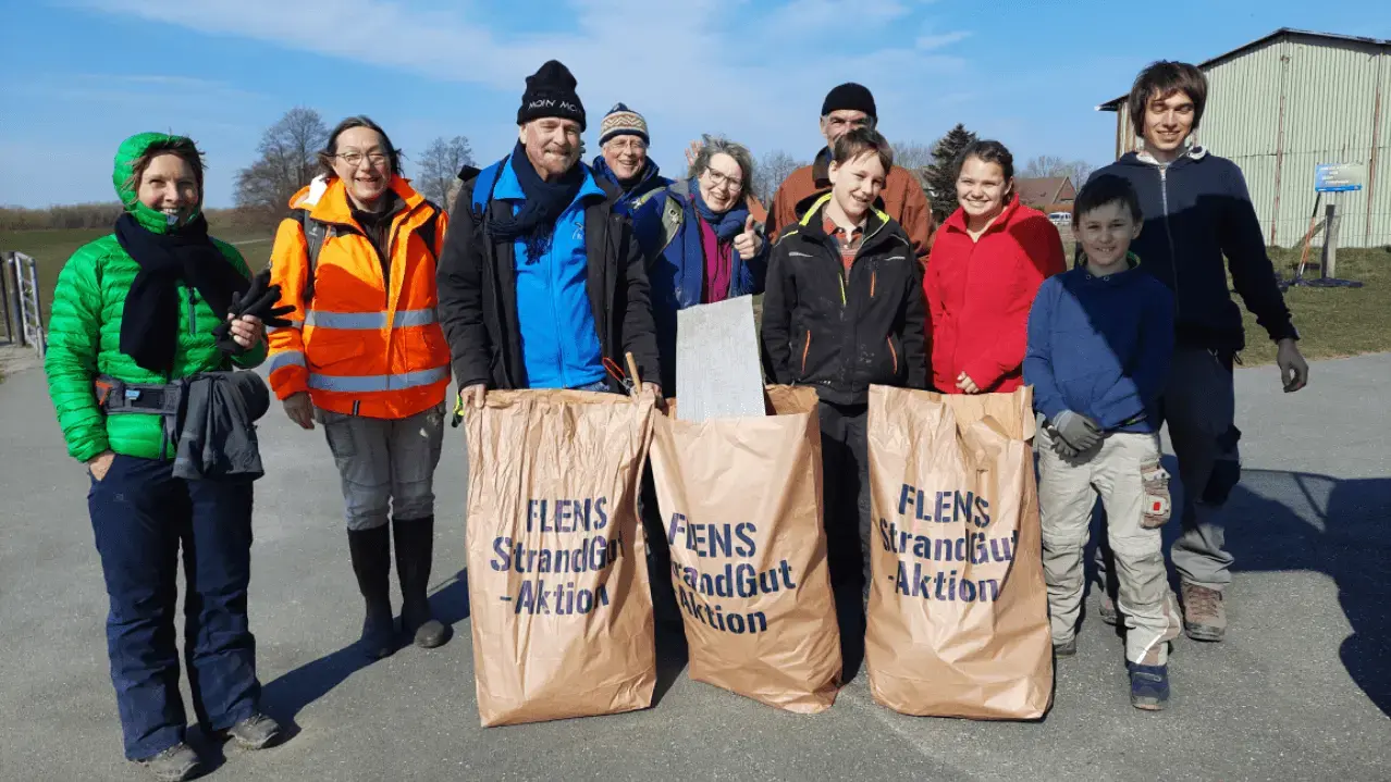 Gruppe von Freiwilligen bei einer FLENS Strandreinigung mit Müllsäcken, sonniger Tag.
