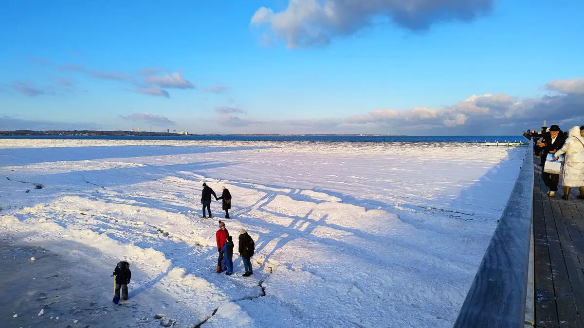 Menschen auf zugefrorenem Strand und Eisfeld neben Seebrücke unter klarem blauen Winterhimmel.