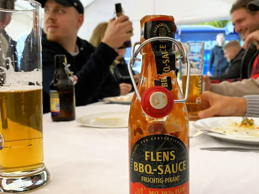 Flensburger BBQ-Sauce auf einem Tisch bei einem geselligen Event im Zelt mit Bier und Personen.