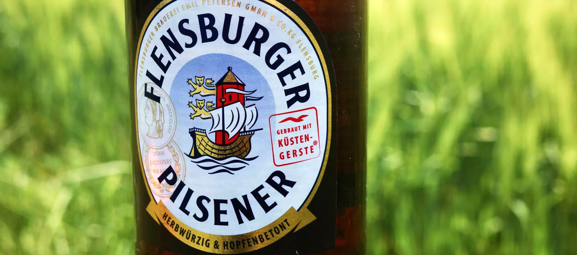 Etikett vom Flensburger Pilsener im Anschnitt vor einer grünen Wiese.