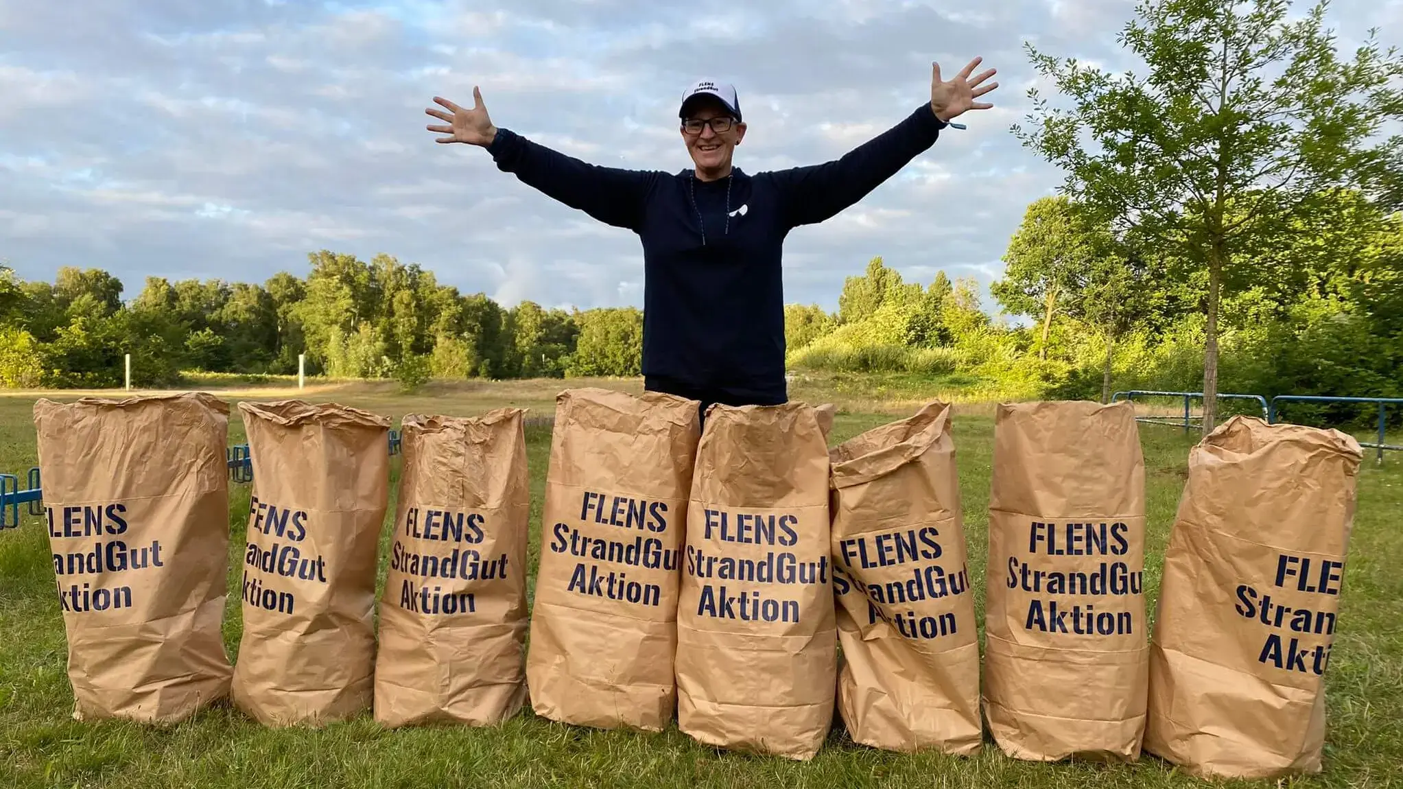 Mann steht zwischen Papiermüllsäcken für Umweltaktion "FLENS StrandGut Aktion" auf einer Wiese.