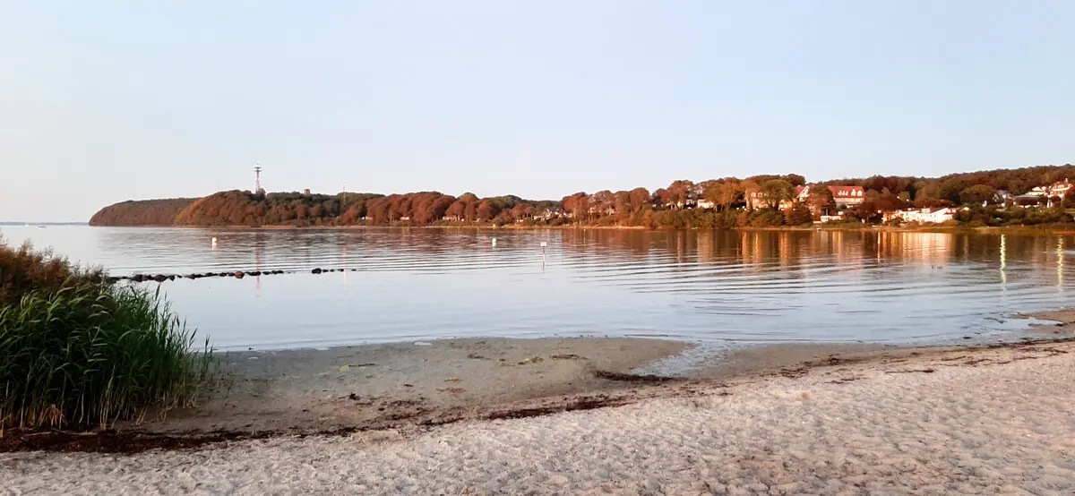 Sandstrand und ruhige Bucht von Solitüde in Flensburg im Abendlicht mit Küstenhäusern und Leuchtturm auf bewaldeter Landspitze.