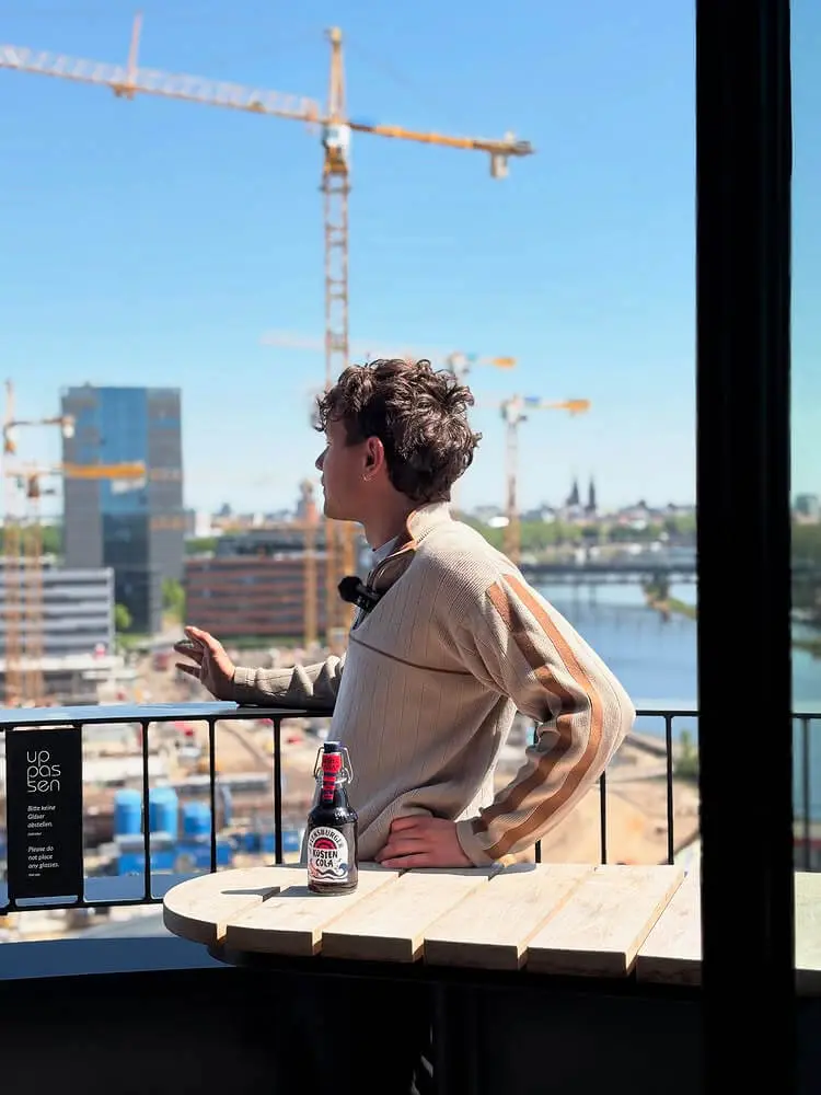 Ein junger Mann führt ein Interview auf einem Balkon vor einer Stadtkulisse, aber dem Tisch eine Flensburger Küstencola.