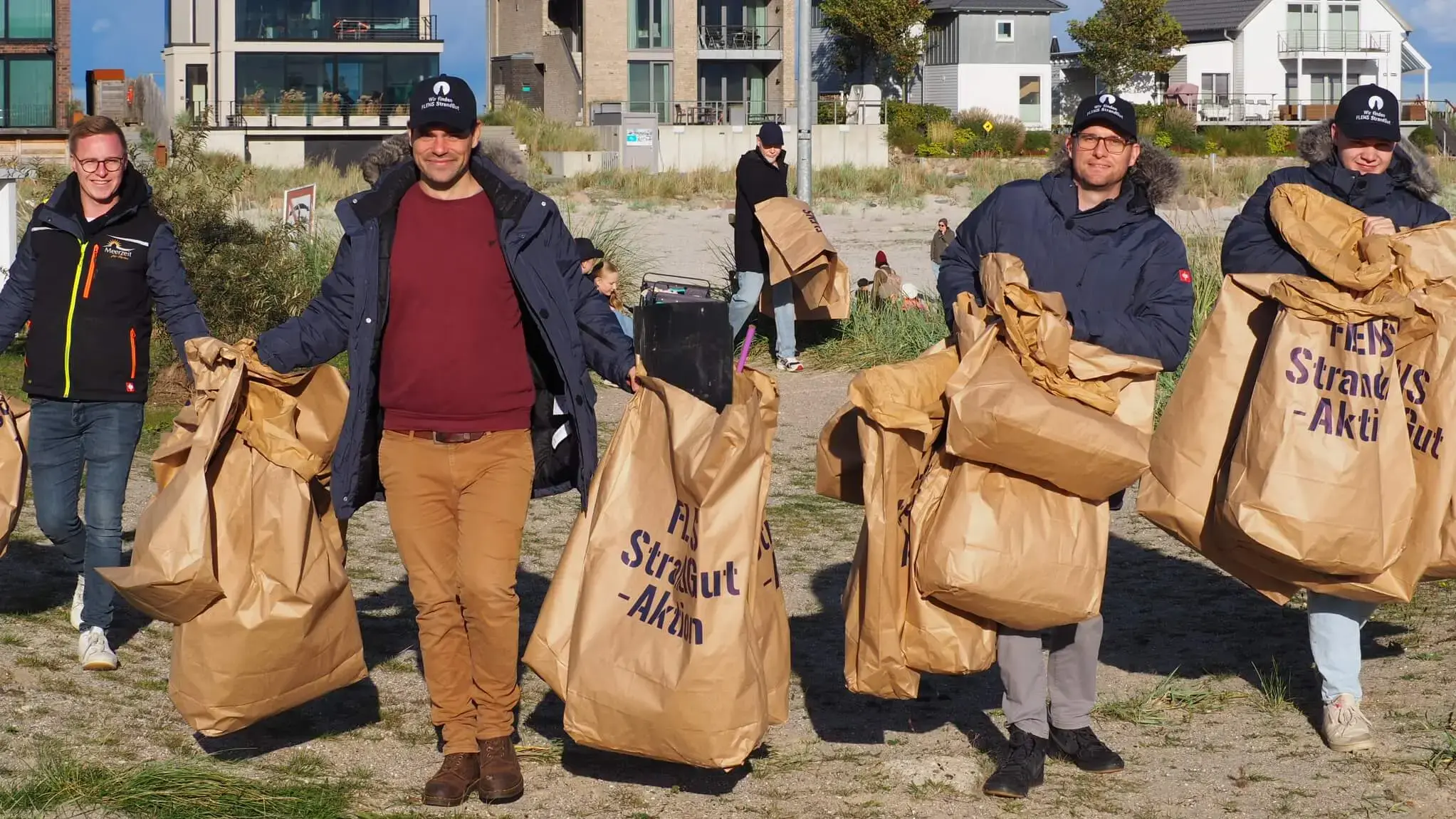 Freiwillige sammeln Müll am Strand mit Säcken für eine FLENS StrandGut Aktion, moderne Wohnhäuser im Hintergrund.