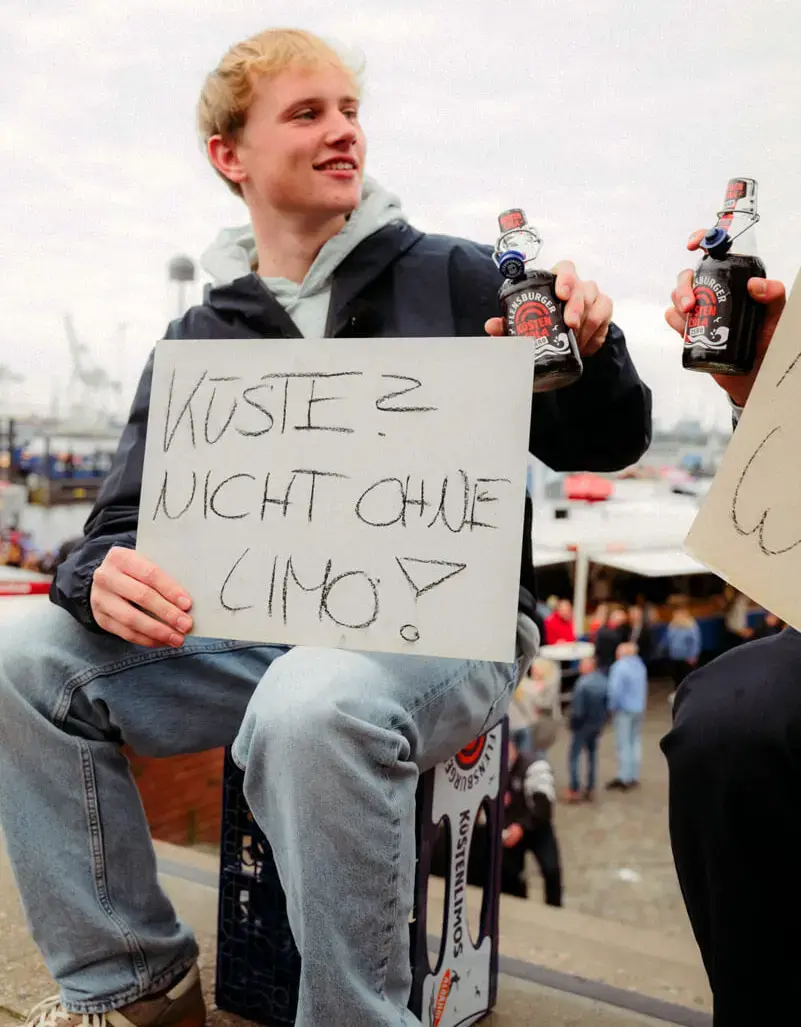 Jugendlicher am Hafen hält Schild "Küste? Nicht ohne Limo!" und Bügelflaschen mit Flensburger Küstencola Zero.