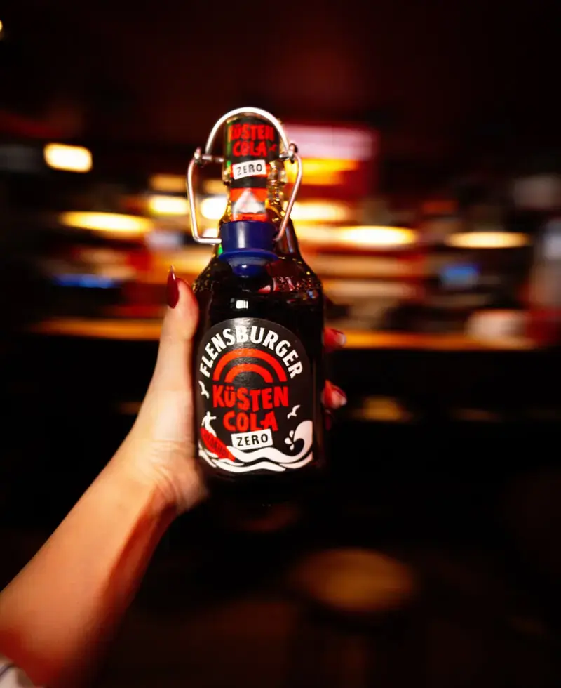 Hand hält Flensburger-Küstencola-Zero-Flasche mit Bügelverschluss vor unscharfem Bar-Hintergrund.
