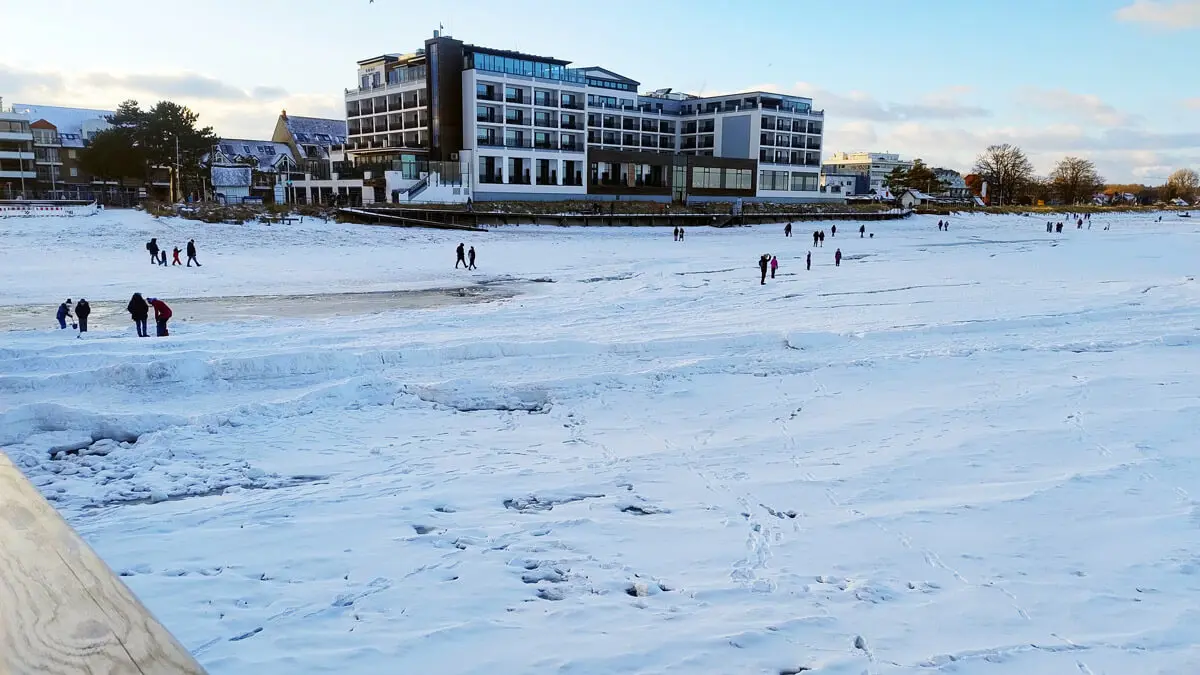 Vereister Strand mit Hotelpromenade, Spaziergängern und Fußspuren im Schnee.