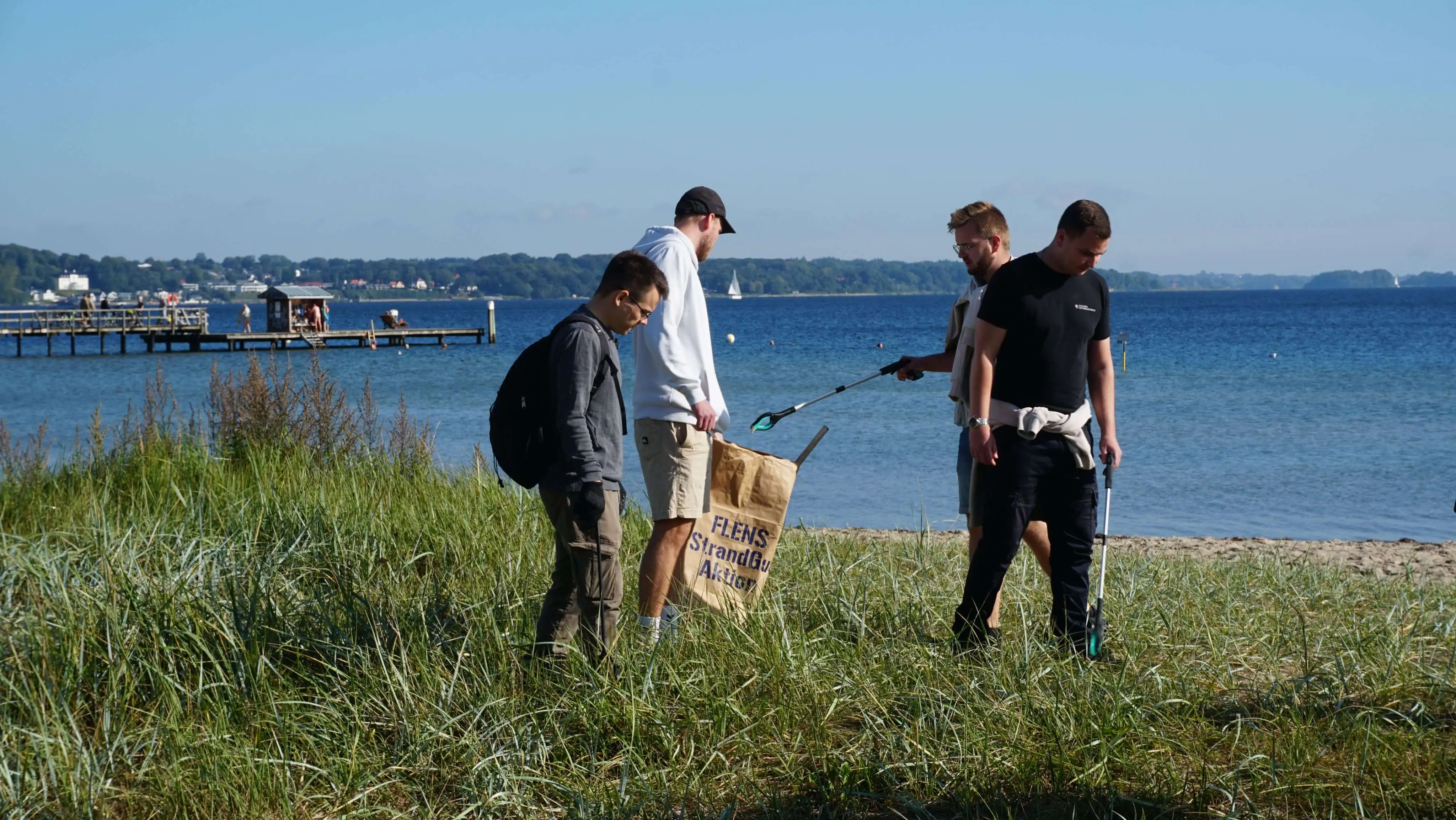 Freiwillige sammeln Müll am Strand bei einer FLENS StrandGut Aktion mit Blick auf eine Seebrücke und Segelboote auf ruhigem Wasser.