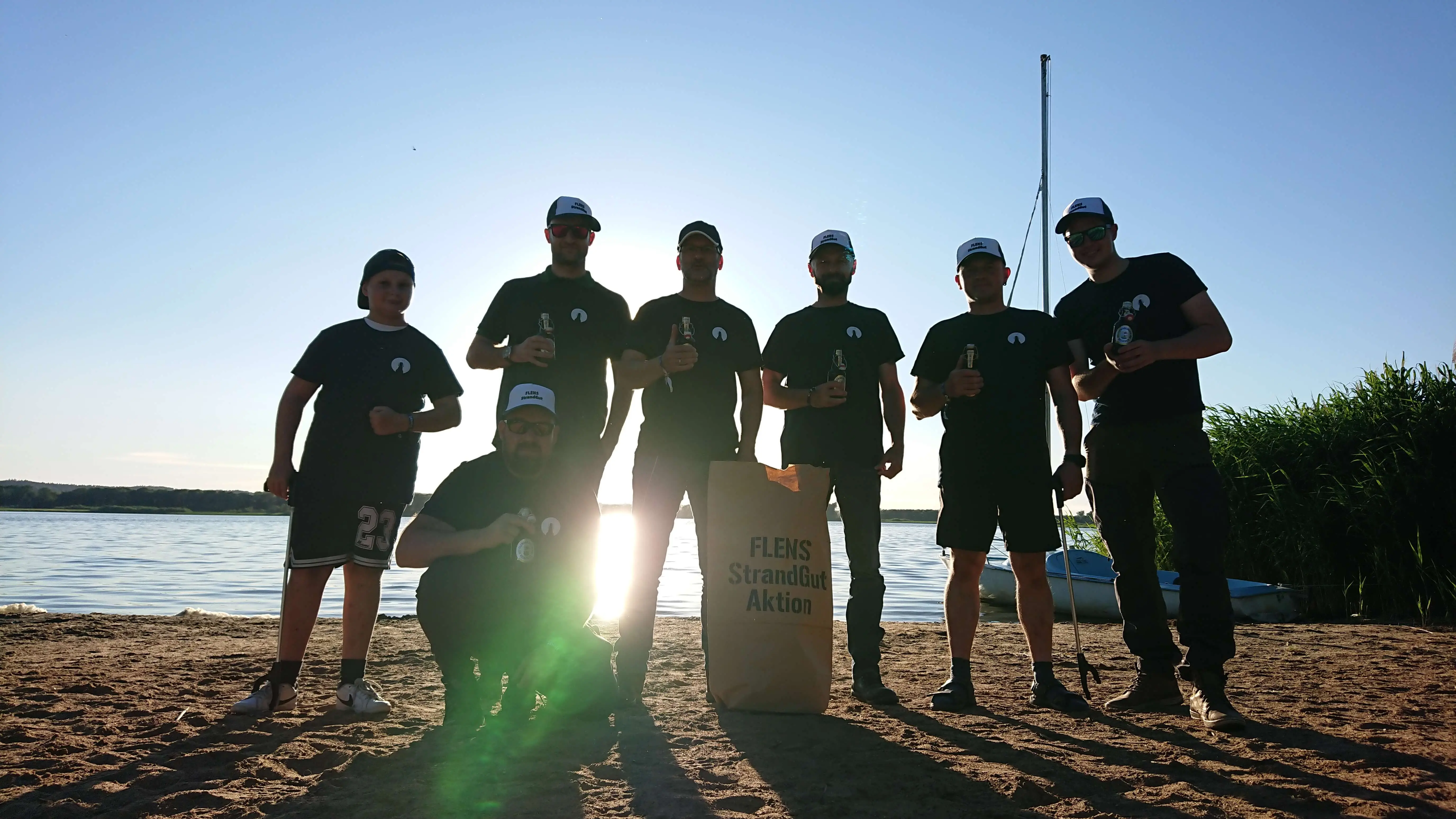 Gruppe junger Männer in einheitlichen FLENS Shirts mit FLENS Flaschen am Strand bei Sonnenuntergang.