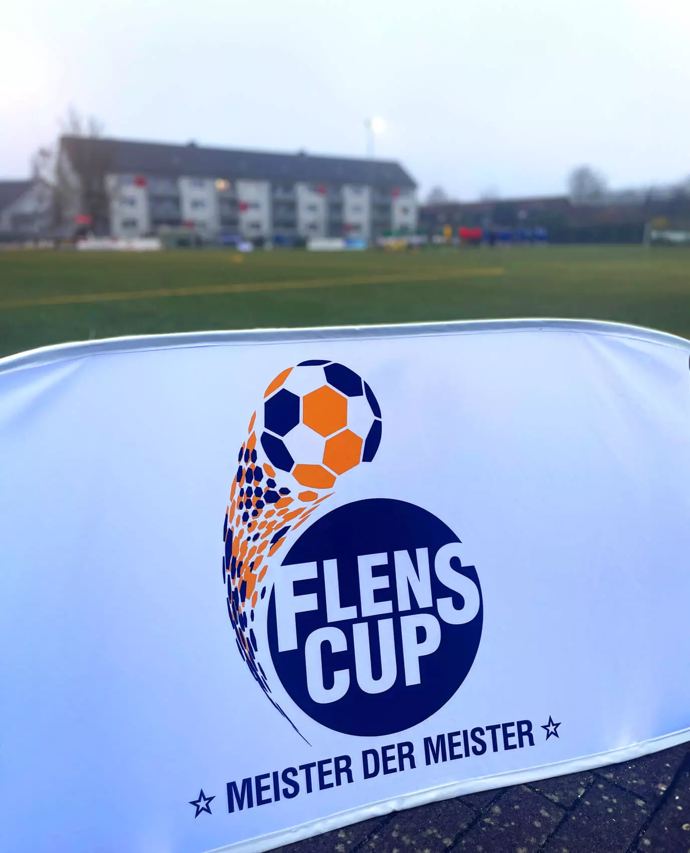 Logo vom FLENS CUP Meister der Meister auf einer Bande vom Fußballfeld.