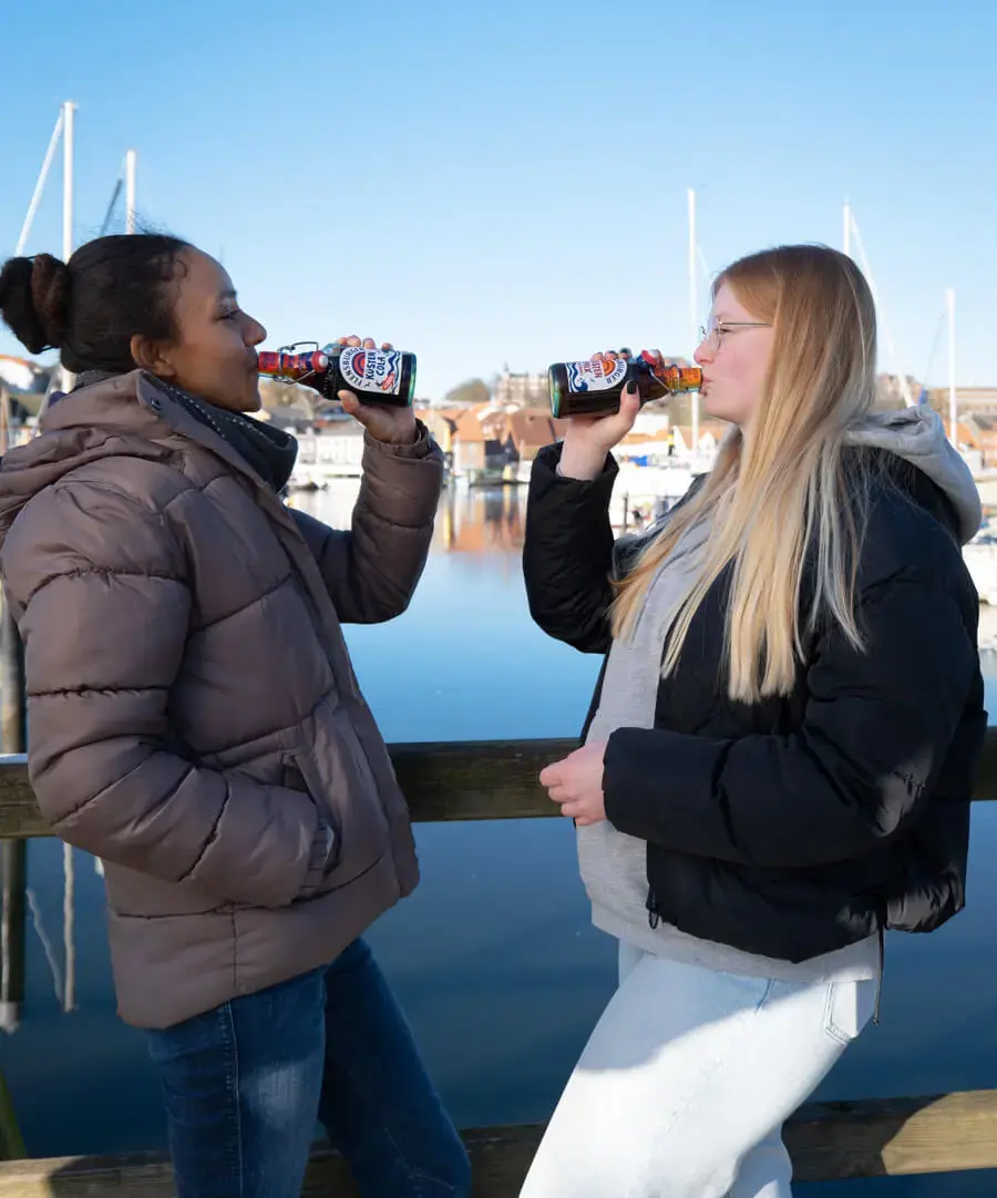 Zwei Freundinnen trinken Flensburger Küstenlimos am Hafen von Flensburg auf Steg, Segelboote im Hintergrund.